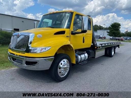 Yellow flatbed tow truck parked on the side of the road with a metal building and trees in the background.
