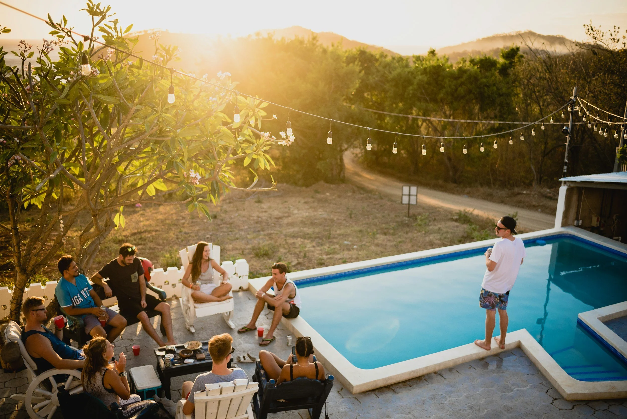 People gathered around a backyard swimming pool with one person standing on the pool's edge, another sitting by the pool, and multiple others sitting on outdoor chairs, enjoying a social gathering during sunset. String lights hang above the scene, and there are trees and a mountain in the background.