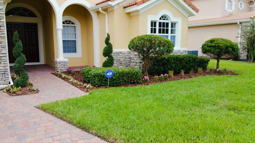 Front yard of a house with a brick pathway, manicured bushes, trimmed trees, and green grass, with an ADT security sign.