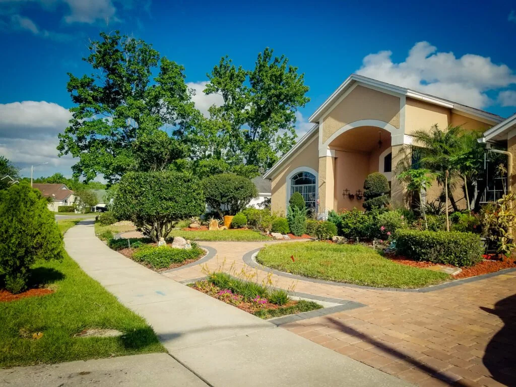 A residential front yard with a paved brick driveway, manicured bushes, and trees, in front of a beige house with an arched entrance and large windows, under a bright blue sky with clouds.