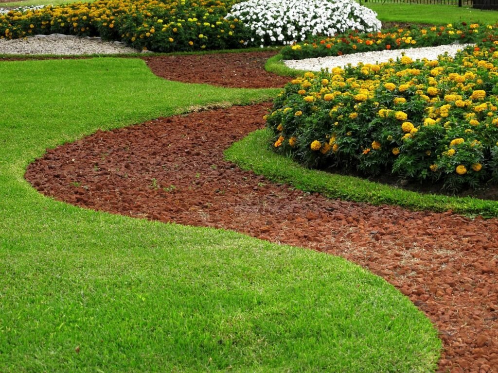 A landscaped garden with a winding red mulch pathway, green grass on both sides, and flower beds with yellow, white, and orange flowers in the background.