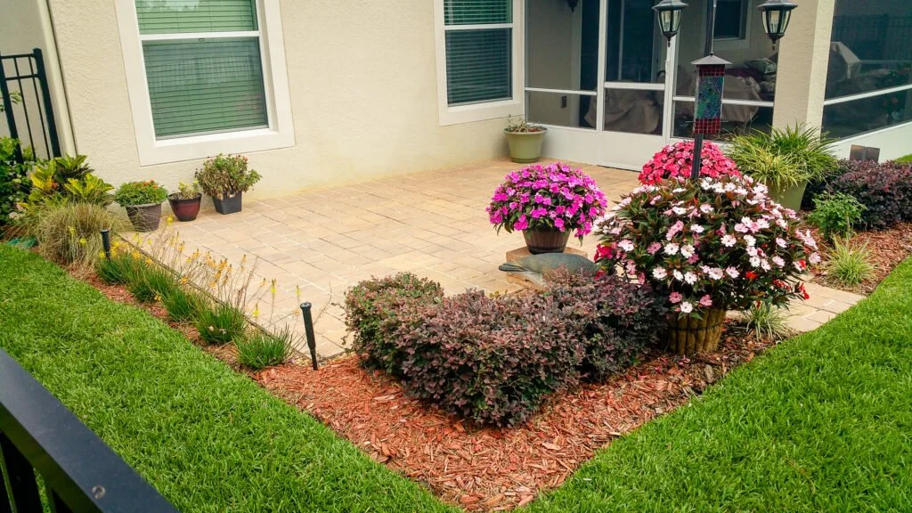 Well-maintained backyard patio with potted and planted flowering plants, a bird feeder, and a screened porch in the background.