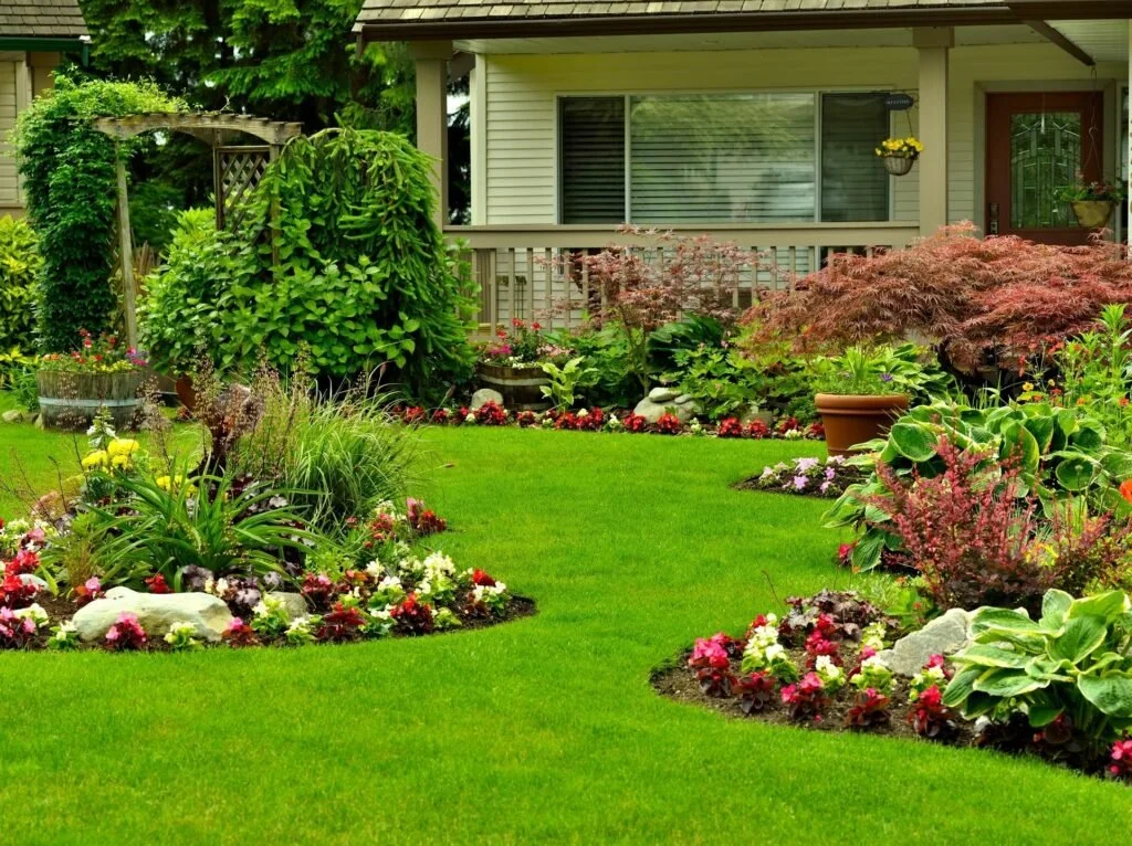A lush garden with well-maintained green grass, flowering plants, shrubs, and small trees in front of a house with a porch. The scene includes decorative rocks, potted plants, and colorful blooms, creating a peaceful outdoor space.
