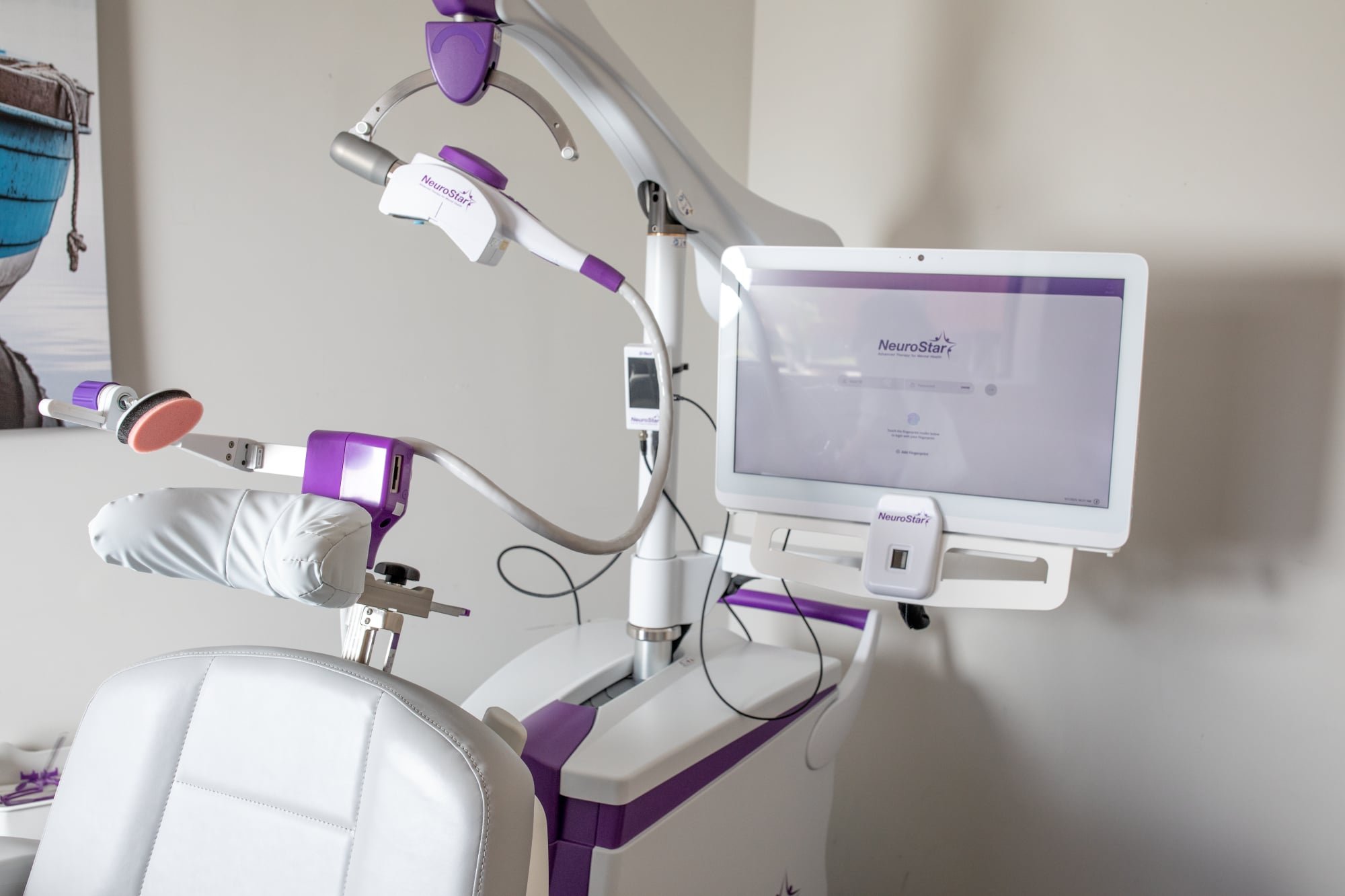 Medical equipment in a clinical setting, including a dental chair, a monitor displaying the NeuroStar logo, and a robotic arm with purple accents, used for neurological or dental treatments.