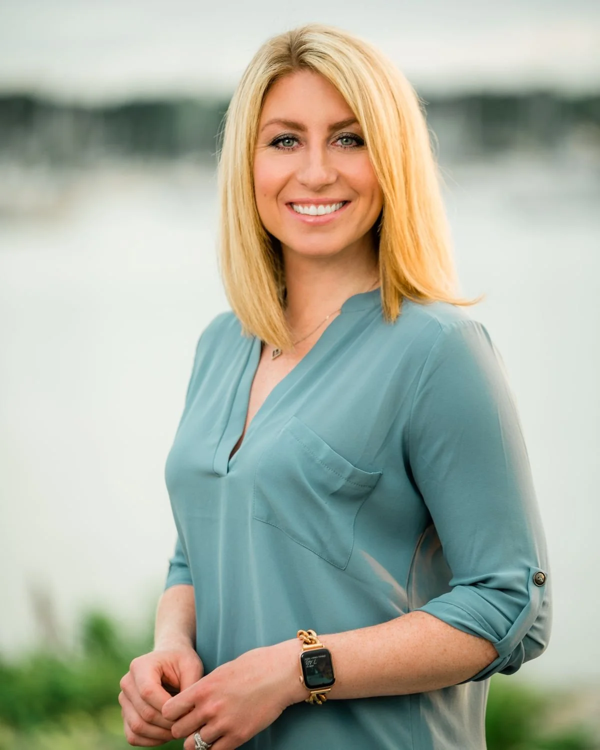 Portrait of a smiling woman with blonde hair wearing a light blue blouse, standing outdoors near a body of water with a blurred background.