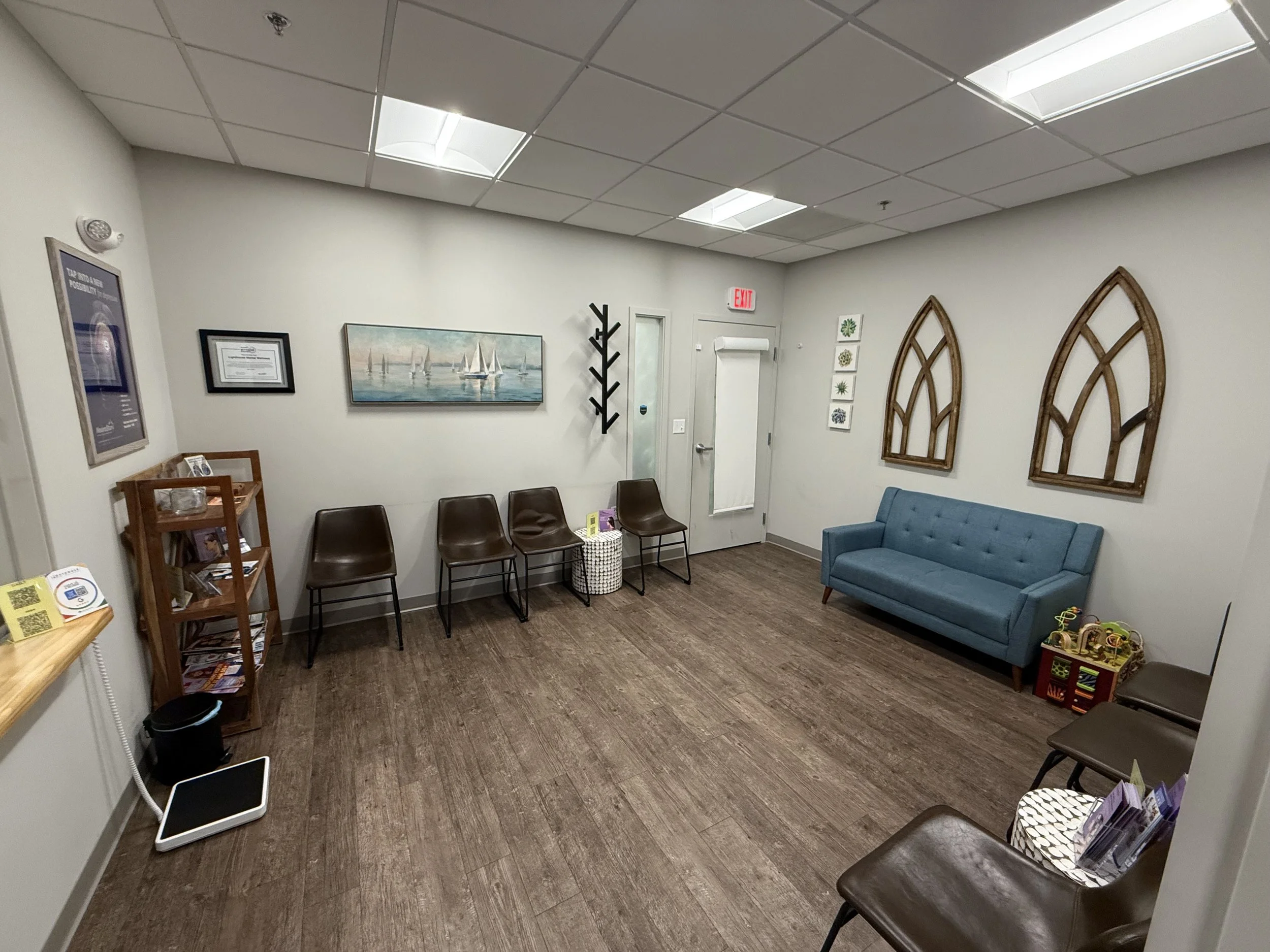 Interior of a waiting room with four black chairs along the wall, a blue sofa, framed artwork, a coat rack, a small table, and children's toys in the corner. The ceiling has white tiles and skylights, and there is an exit sign above a door.