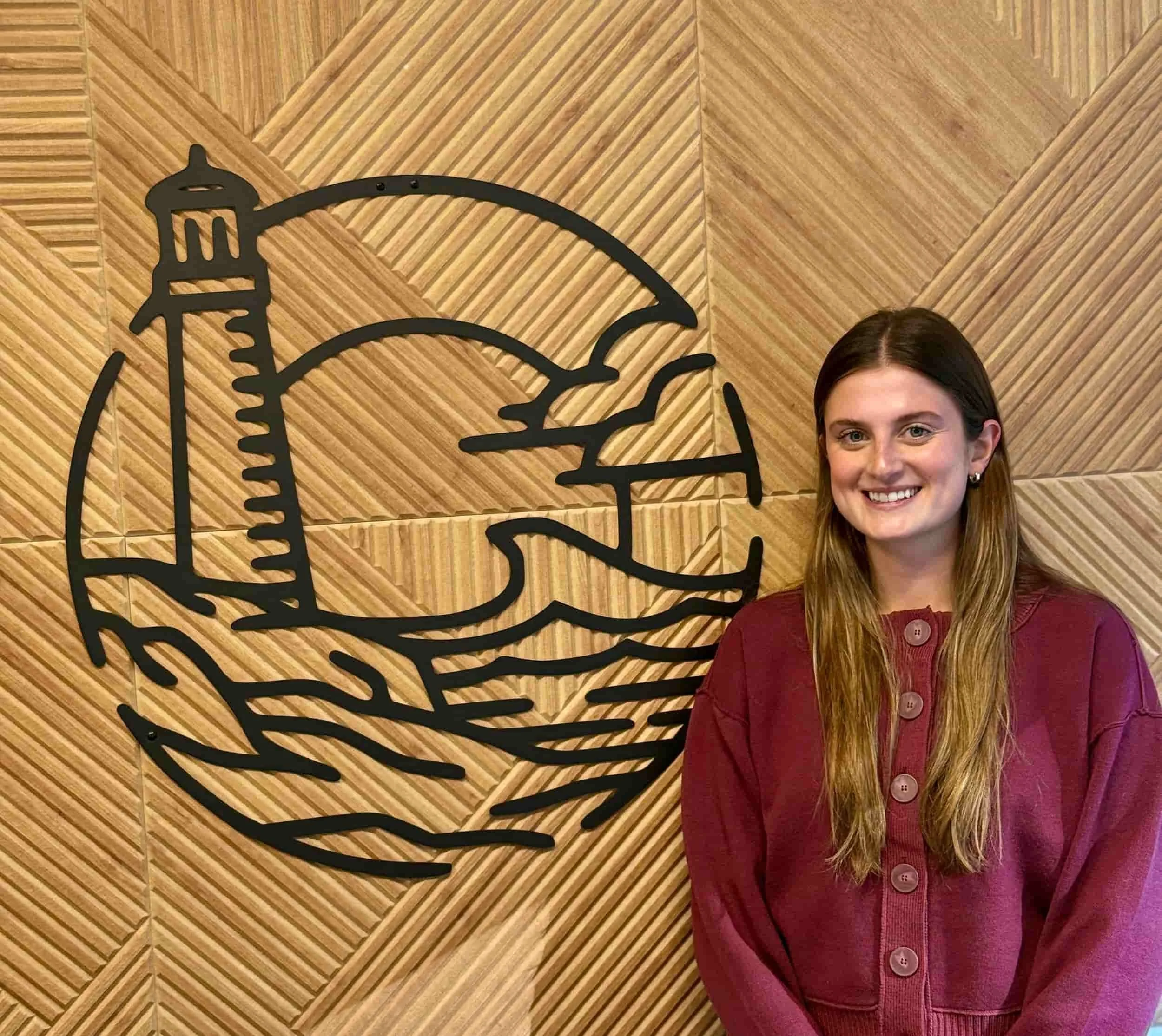 Portrait of a smiling young woman with straight brown hair wearing a maroon sweater, standing in front of a wooden decorative background with black metal outline art.
