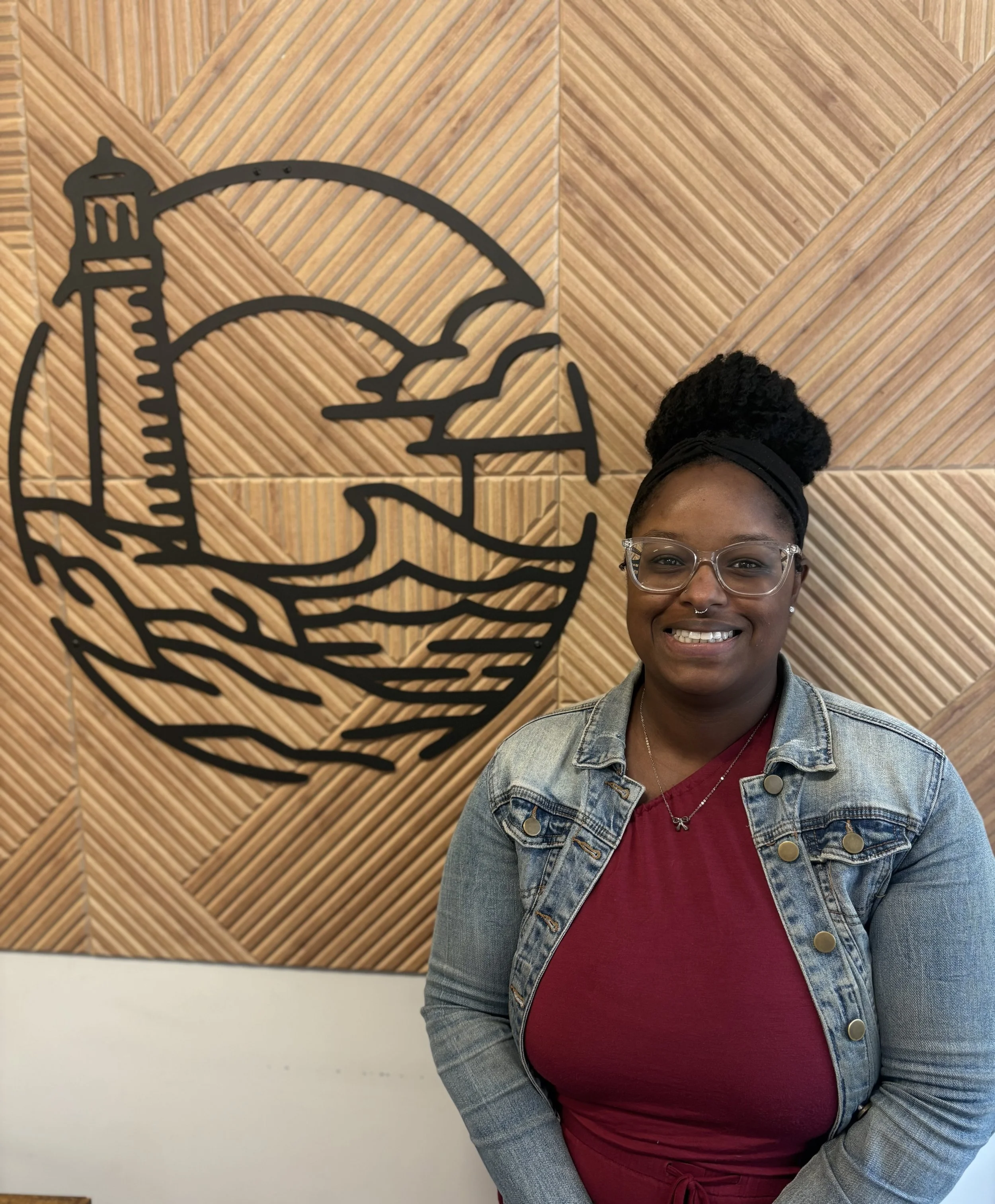 A woman with glasses and a pink shirt wearing a denim jacket, standing in front of a wall with a large black outline of a lighthouse and wave. She has a smile and short curly hair.