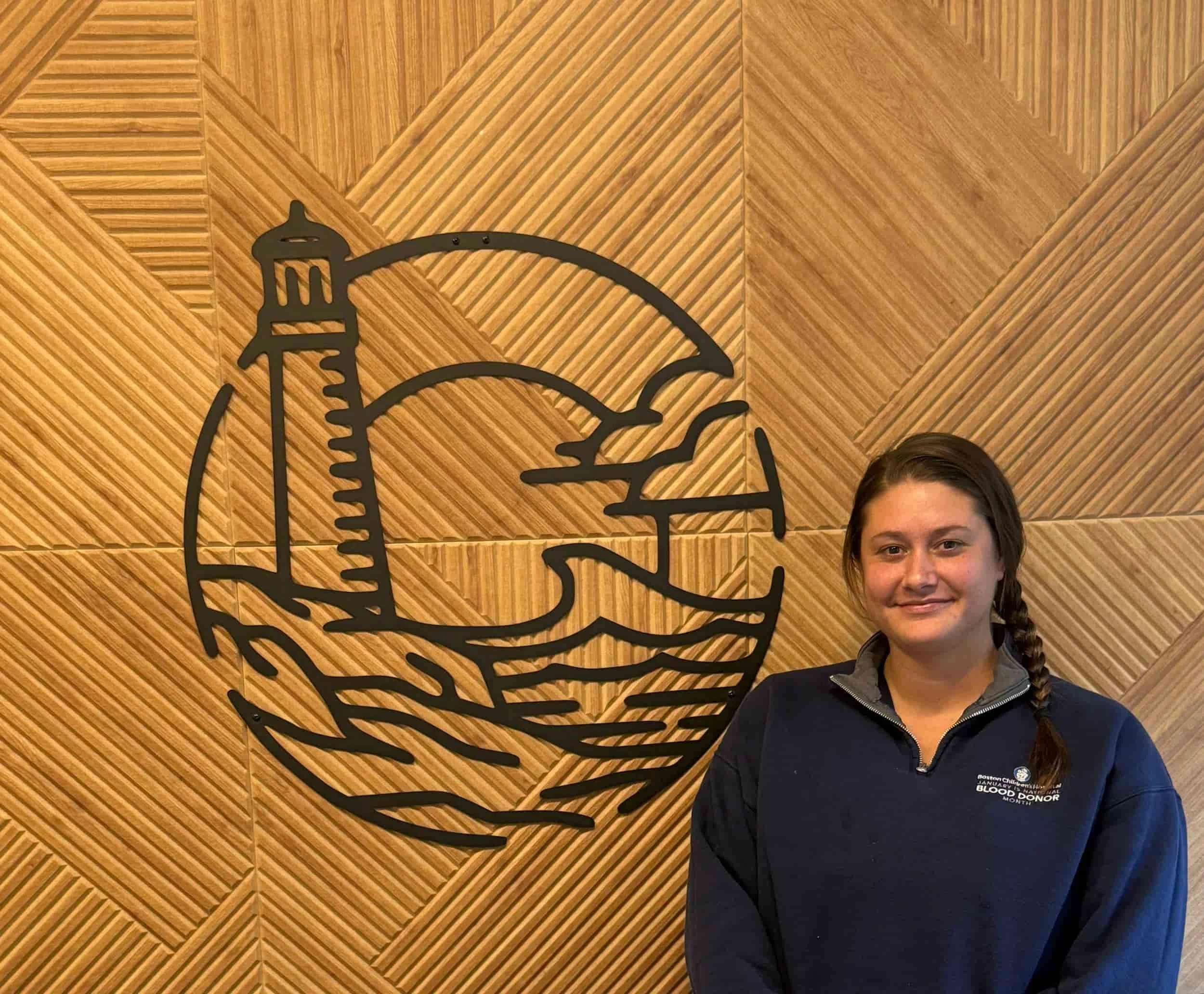 A woman with braided hair and wearing a black sweatshirt standing in front of a wooden wall with a black metal outline of a coffee cup and a bird.