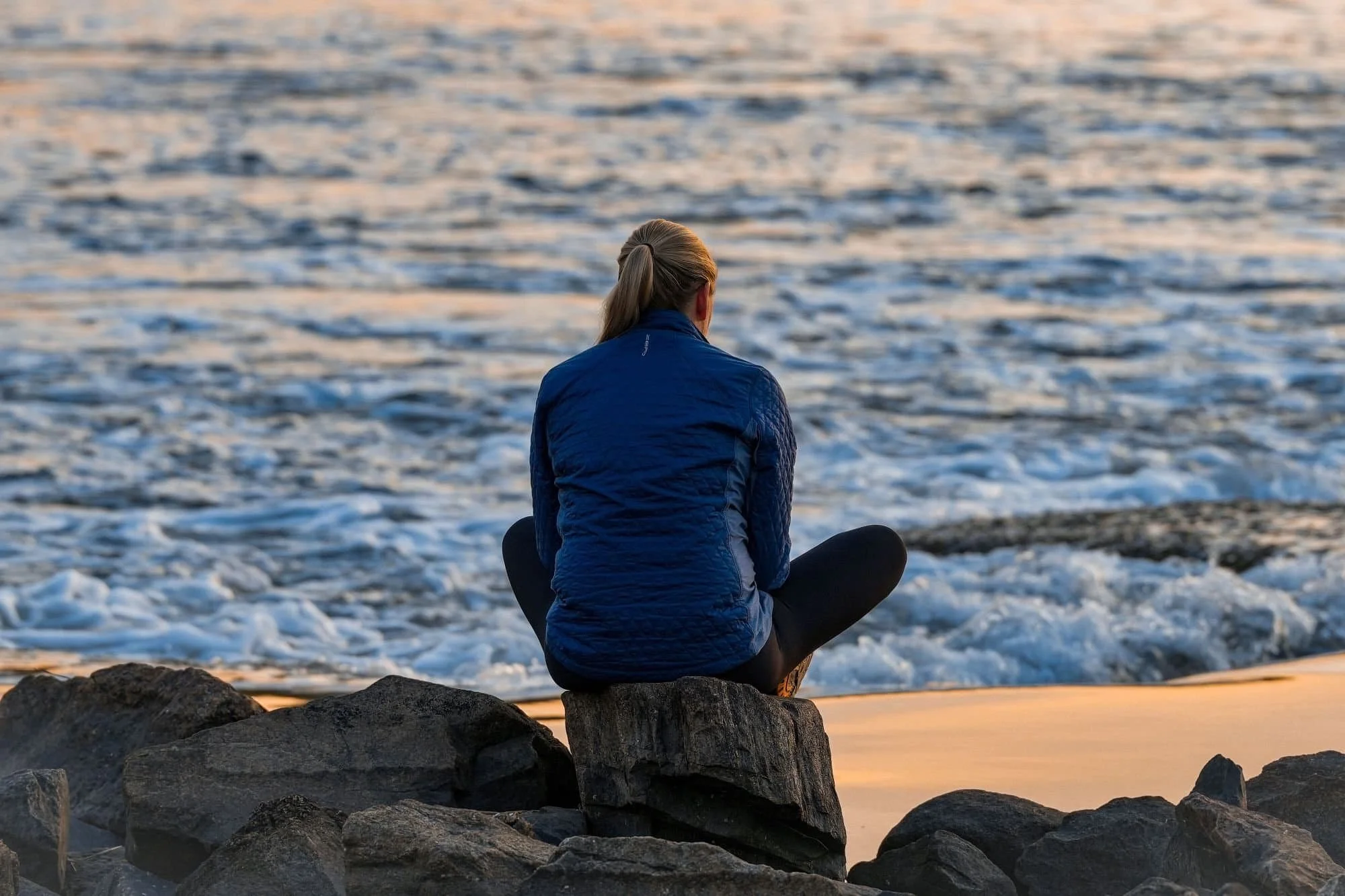 A person sitting on rocks by the water, facing away, with the ocean in the background during sunset or sunrise.