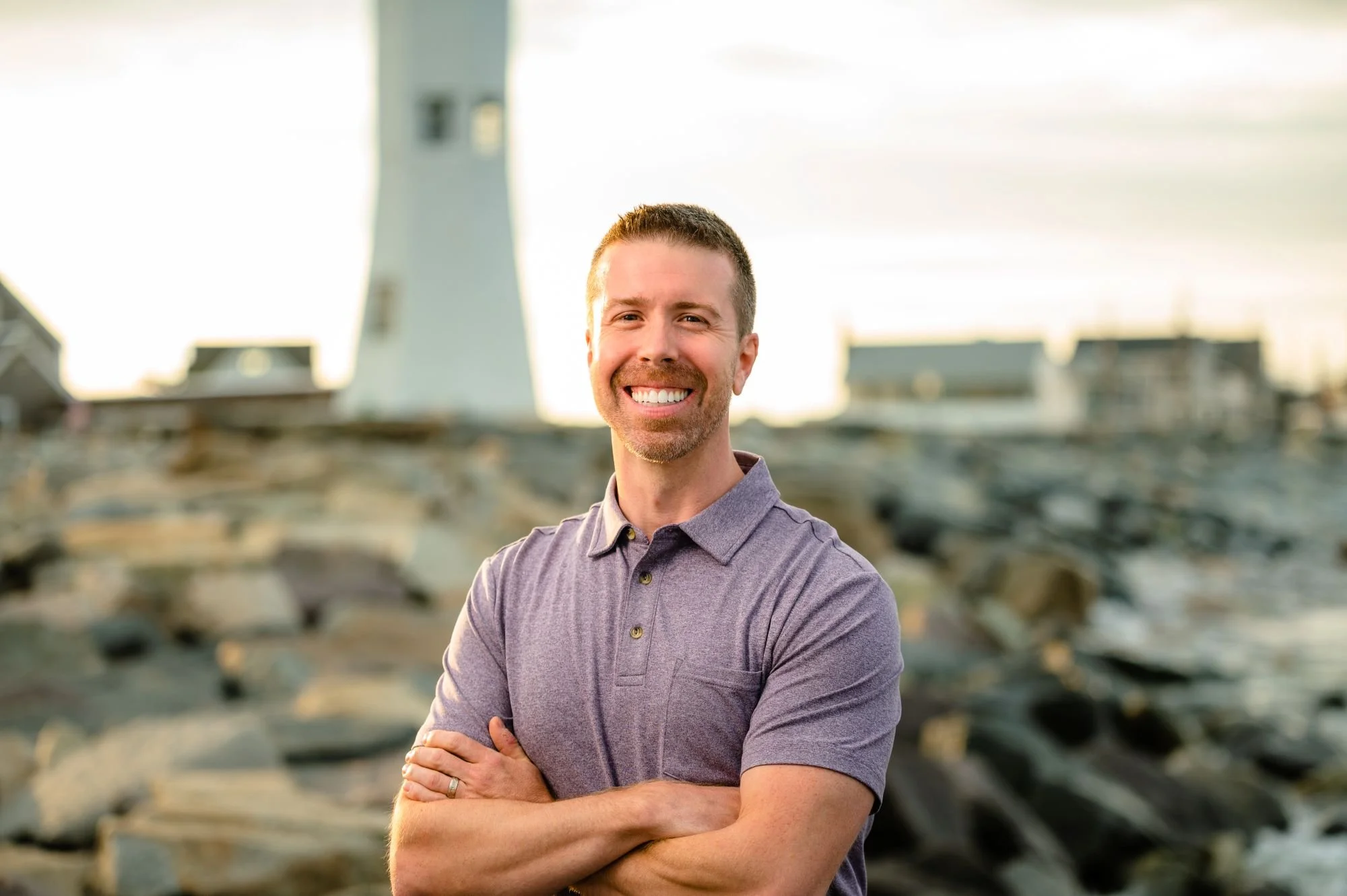 A smiling man with short brown hair and a beard, wearing a purple polo shirt, stands outdoors with arms crossed, in front of a blurred rocky landscape with a lighthouse in the background during daylight.
