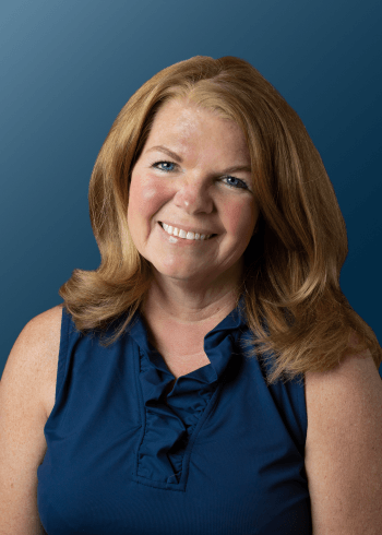 A smiling woman with red hair in a blue sleeveless top against a dark blue background.