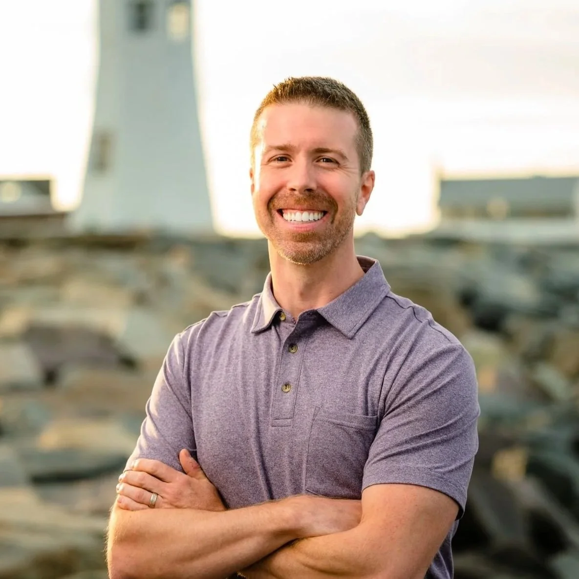 A man smiling with arms crossed, wearing a gray polo shirt, outdoors near a lighthouse.