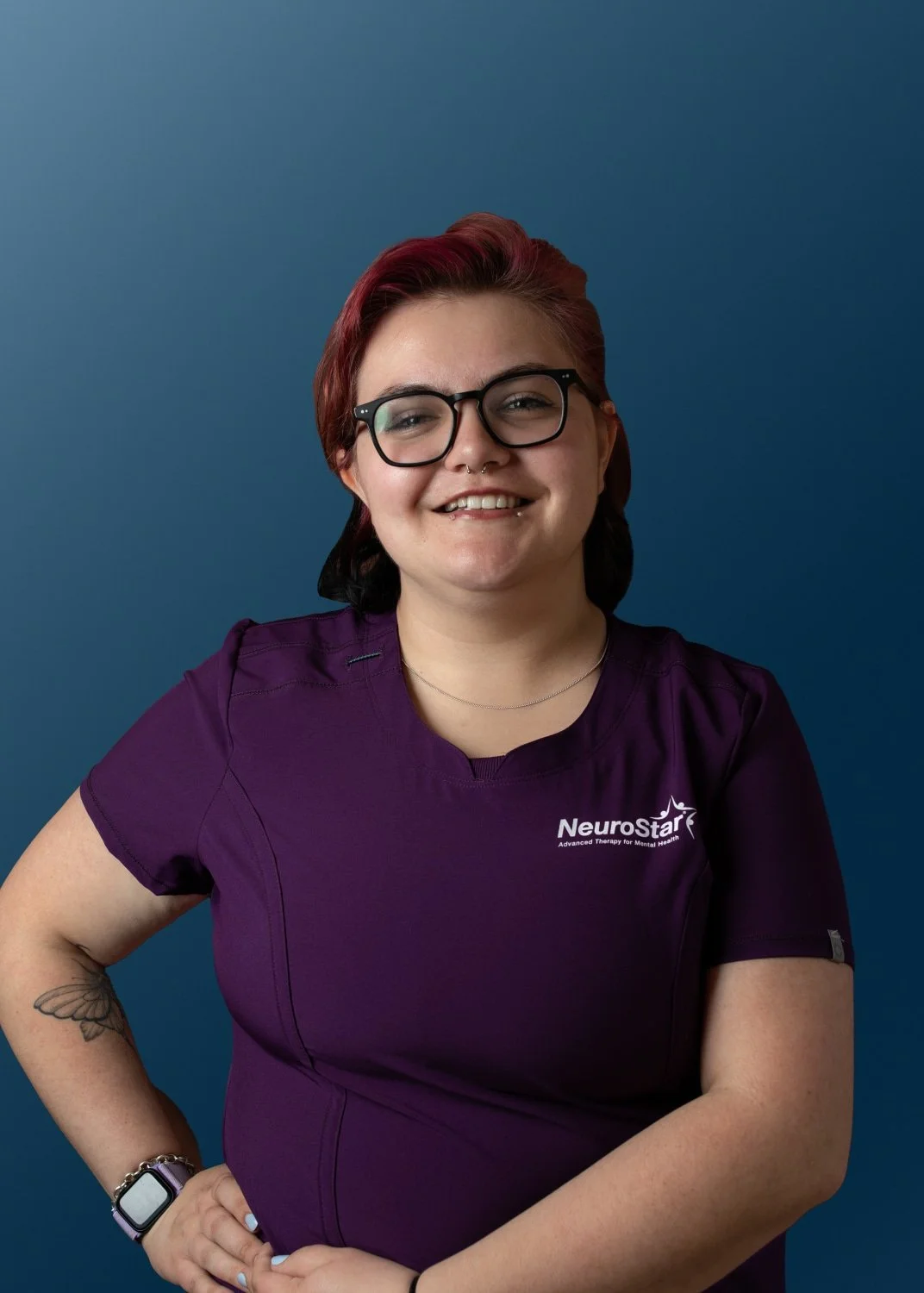 Smiling woman with glasses and short red hair wearing a purple NeuroStar uniform standing against a blue background.