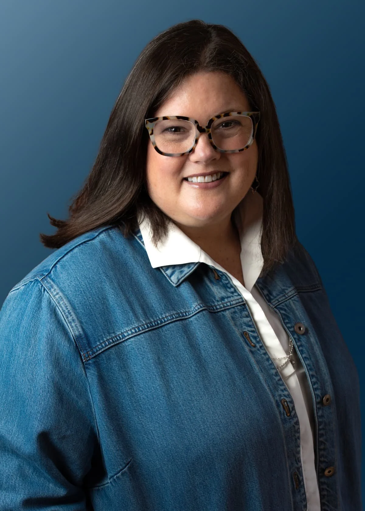 Portrait of a woman with long dark hair, wearing large tortoiseshell glasses, a denim jacket, and a white shirt, smiling against a blue background.