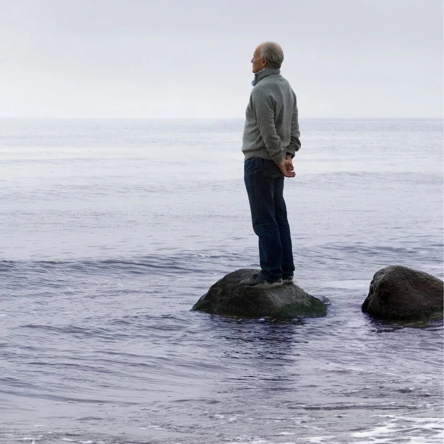 An elderly man standing on a rock in the ocean, looking at the horizon on a cloudy day.