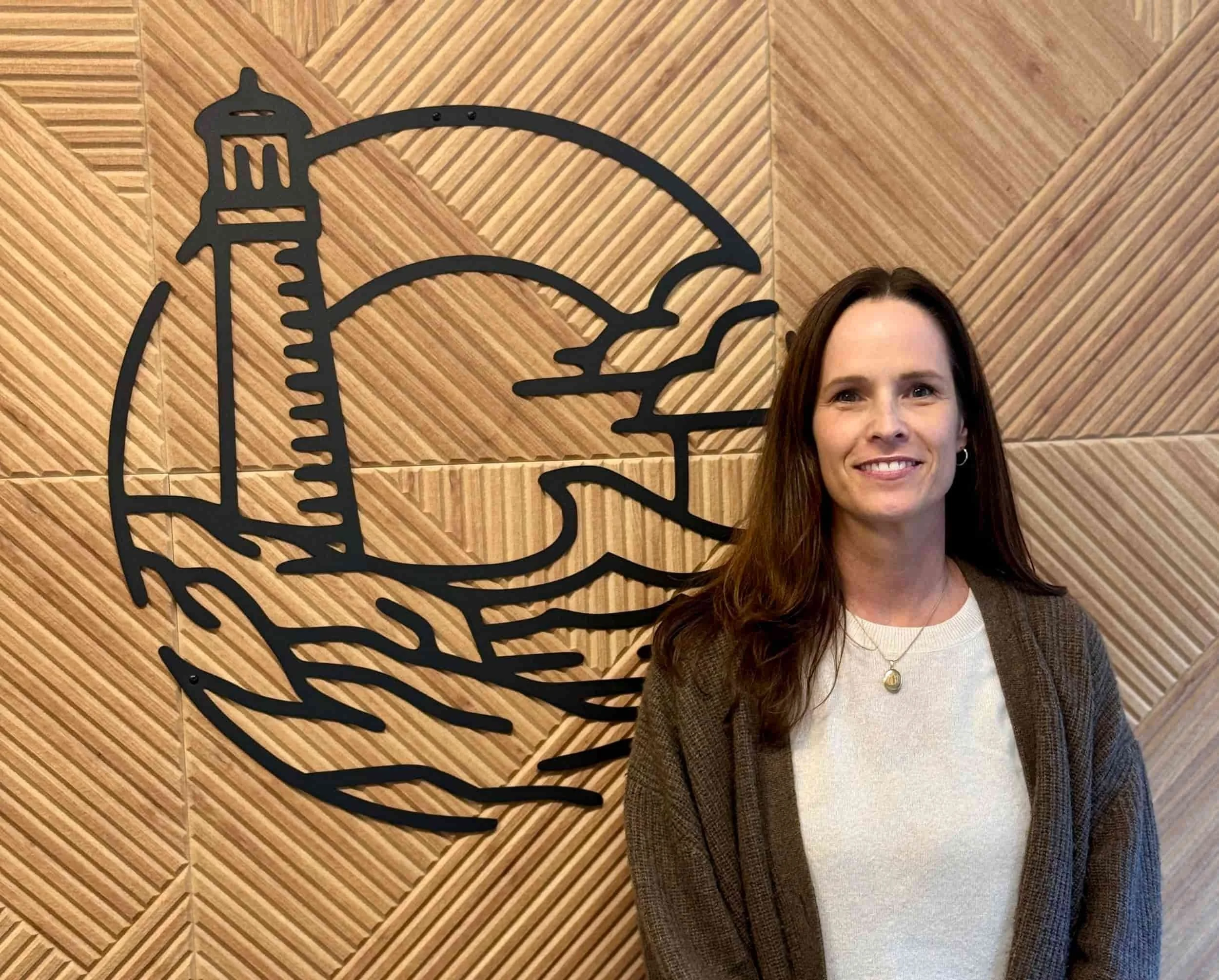 Woman with long brown hair standing in front of a wooden wall with a large black outline wall art of a lighthouse and waves.