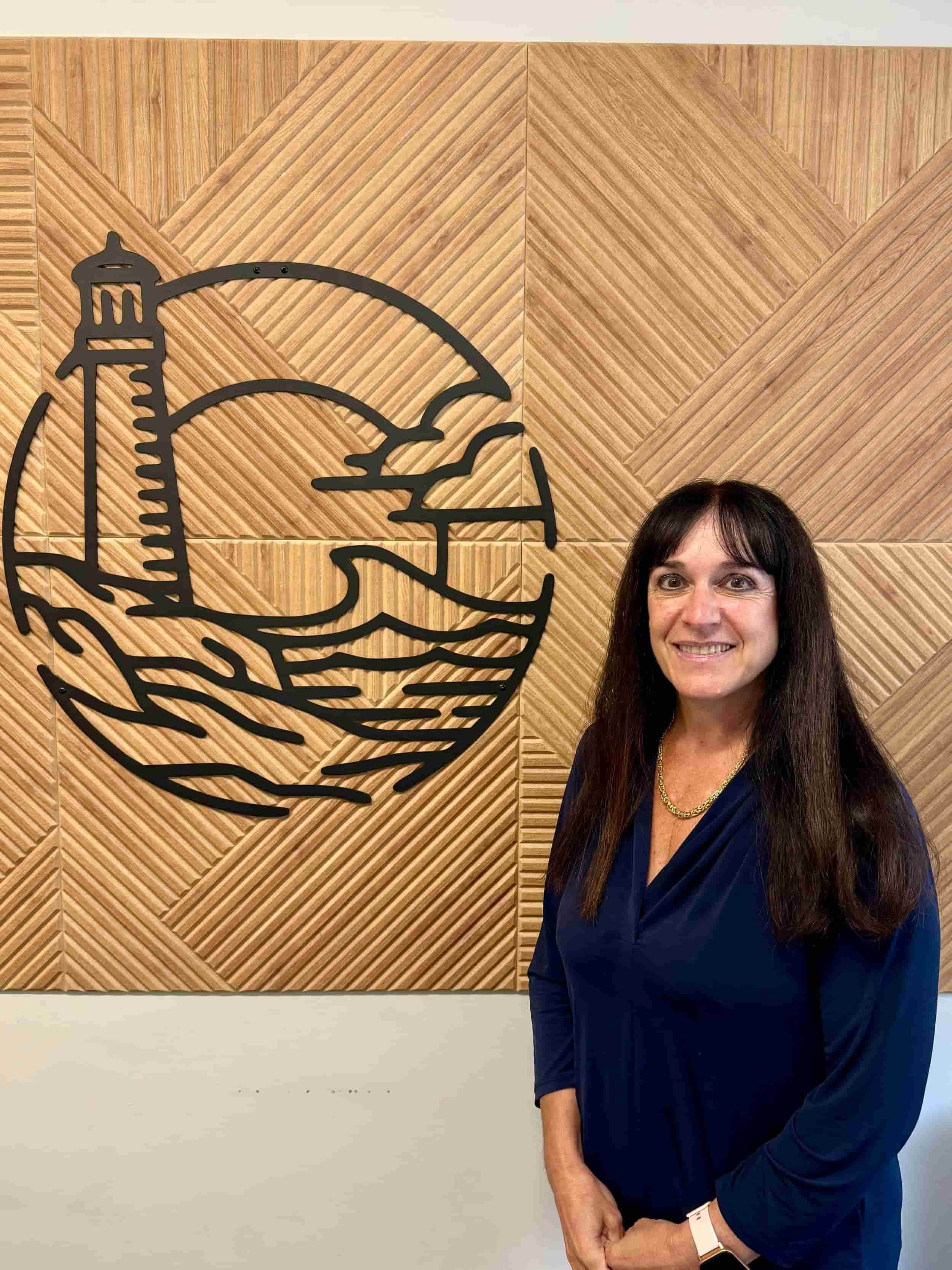 A smiling woman with long dark hair, wearing a navy blue dress and a gold necklace, stands in front of a wooden wall with a black metal art piece. The art piece depicts a lighthouse, a seagull, and a boat on water.