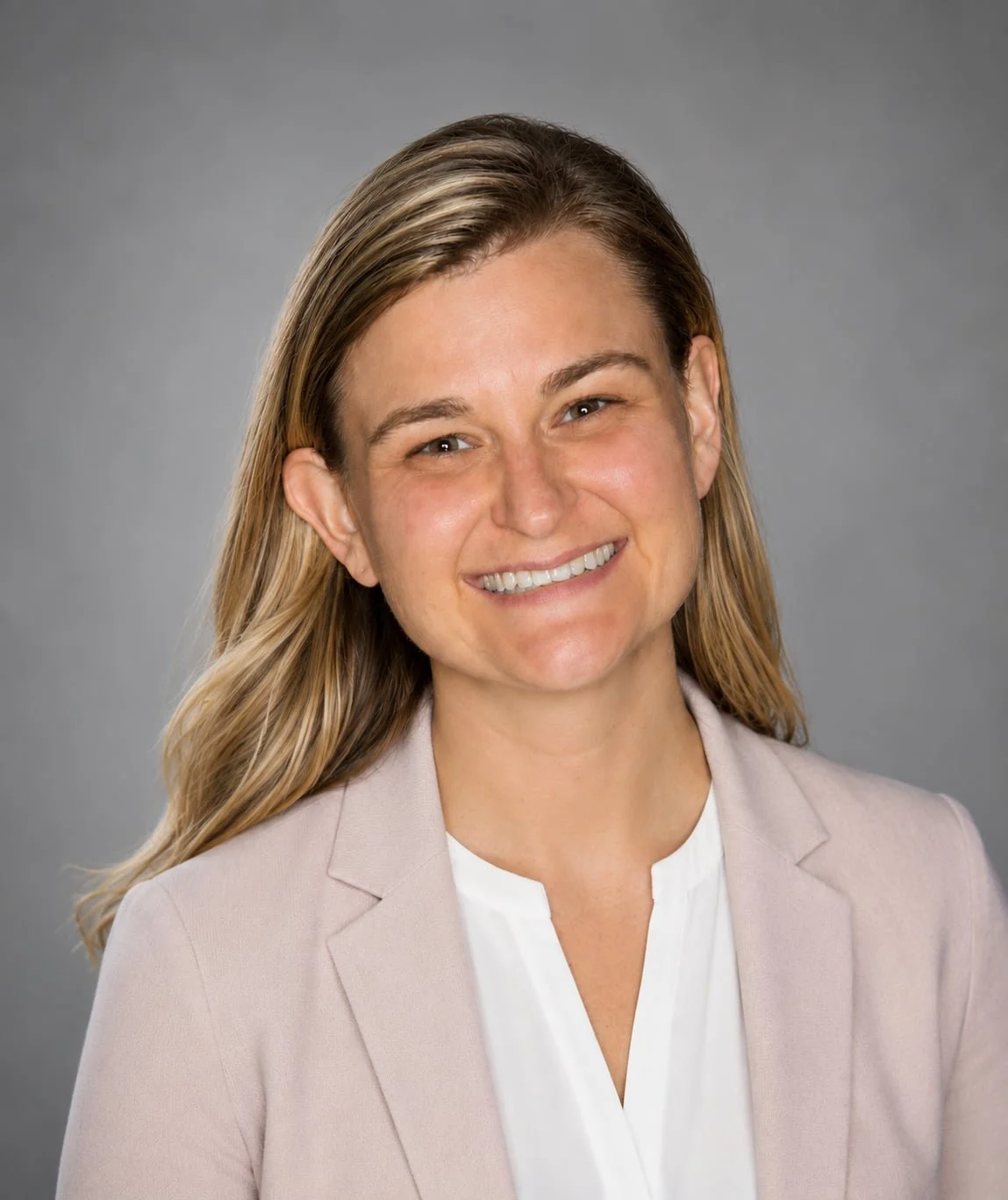 Professional headshot of a woman with blonde hair, smiling, wearing a light blazer and white blouse against a gray background.