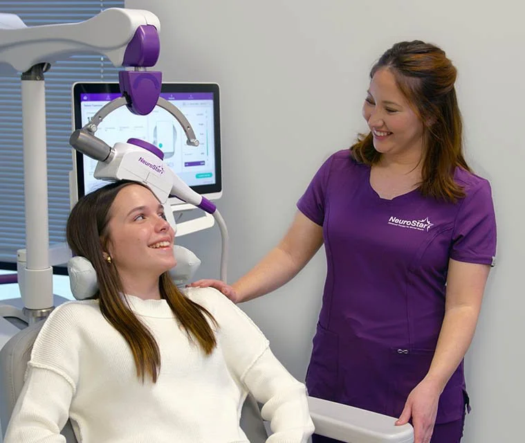 A young woman receiving a brain scan at a medical clinic, smiling and talking with a healthcare professional dressed in purple scrubs.