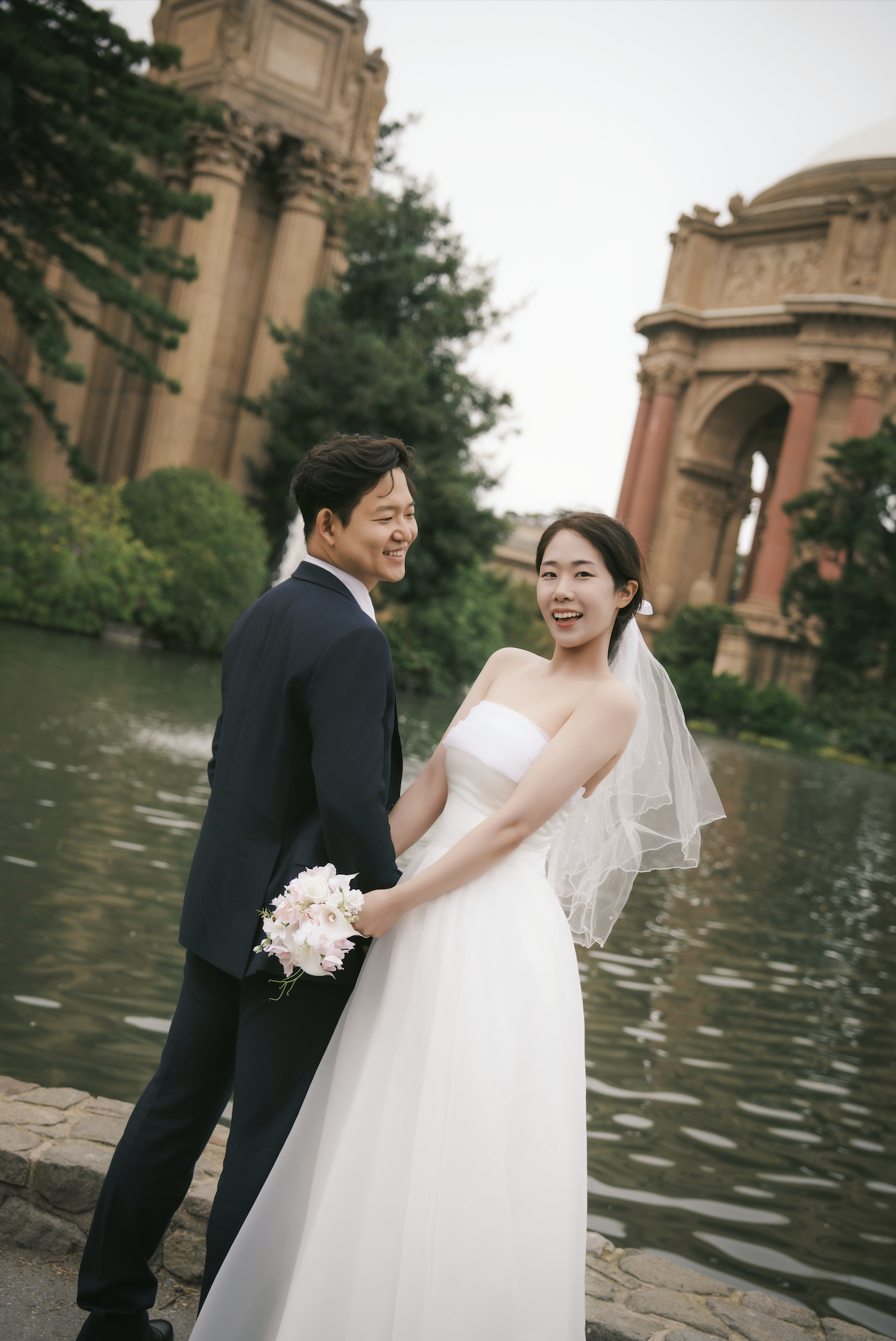 A smiling couple stands near a river, with historic buildings and greenery in the background. The woman holds a bouquet, and the man wears a suit.