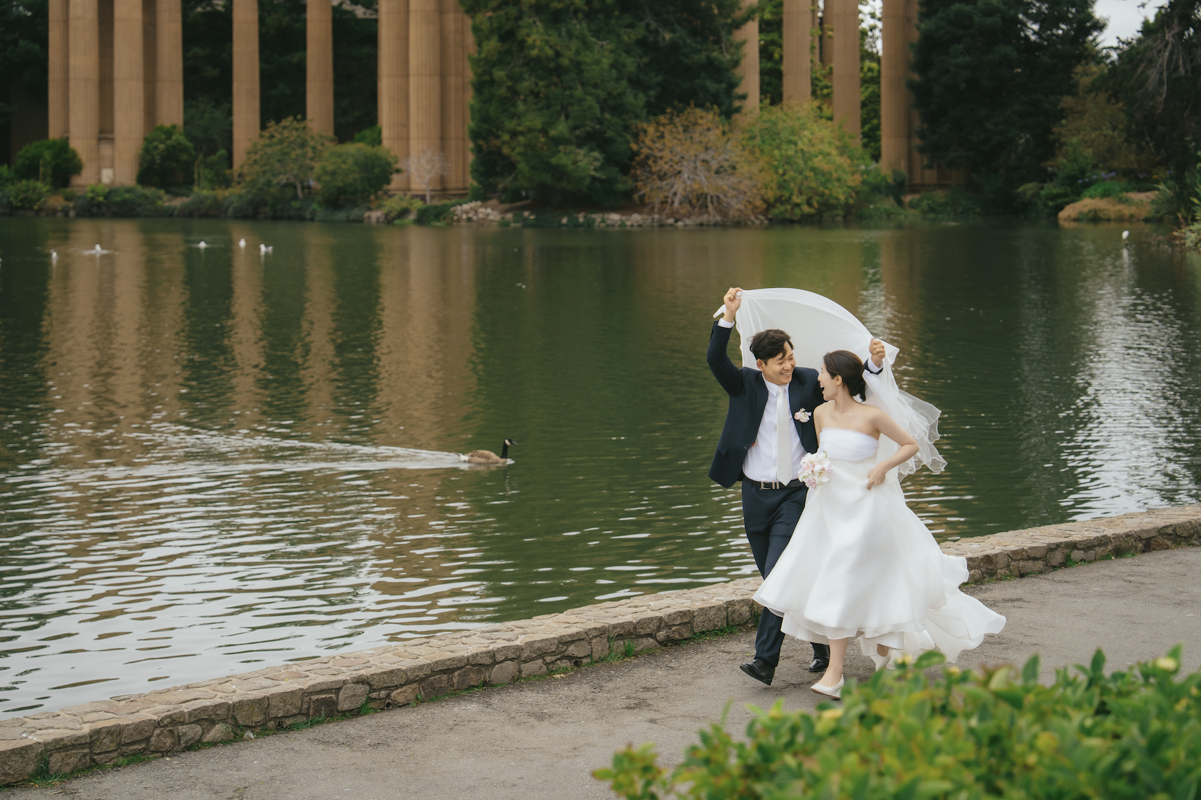 A bride and groom in wedding attire dancing and laughing near a lake with ducks, trees, and tall historical columns in the background.
