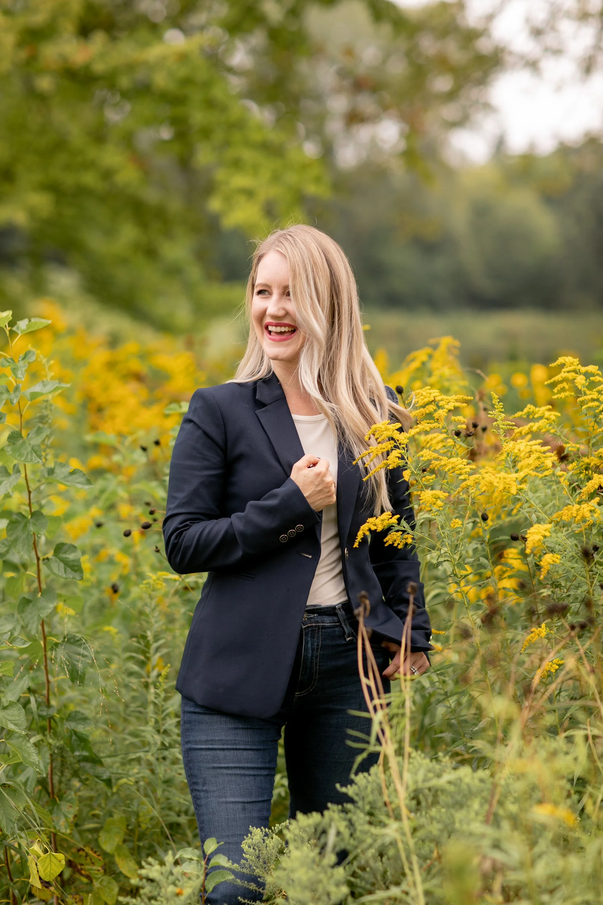 A woman with long blonde hair is smiling and standing in a field of yellow flowers with green trees in the background. She is wearing a dark blazer, a white top, and jeans.