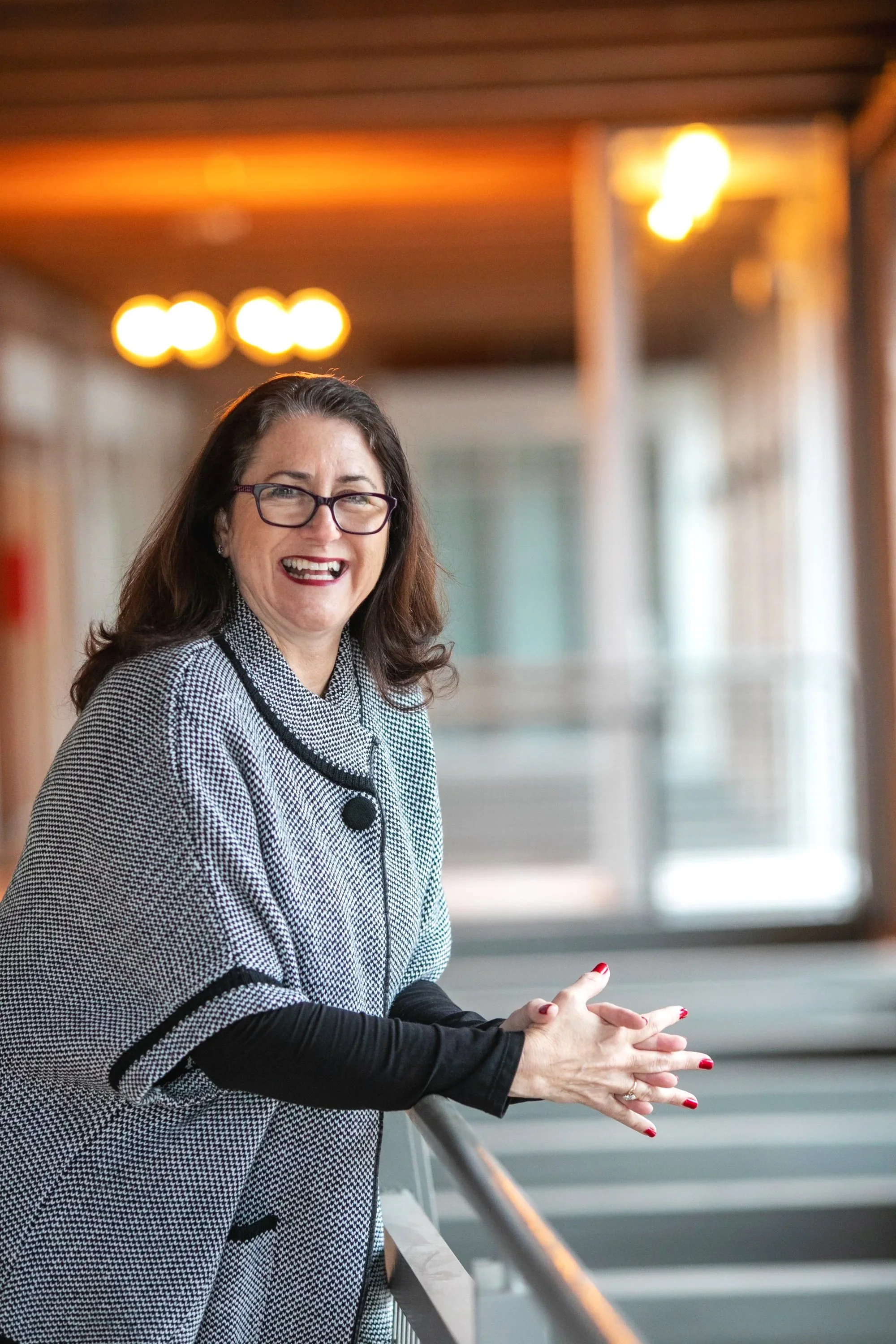 A woman with long brown hair, wearing glasses and a black and white houndstooth blazer, is smiling and holding onto a railing with both hands. She has red nail polish. The background shows an indoor setting with warm lighting and blurred architectural elements.
