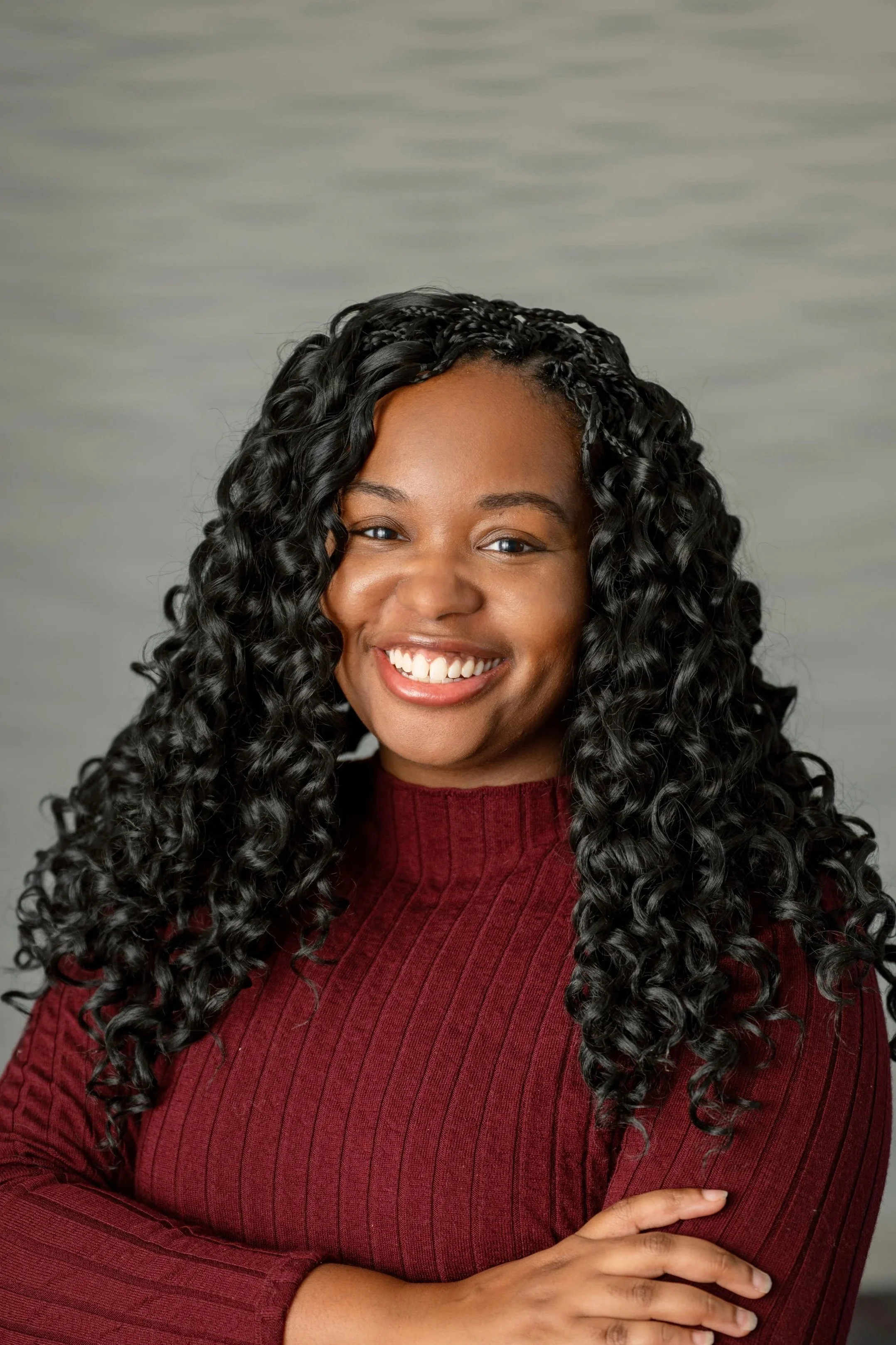 A woman with long, curly black hair wearing a red ribbed turtleneck sweater, smiling with arms crossed, in front of a gray background.