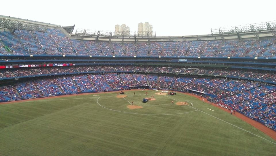 View of a nearly empty baseball stadium with some cleanup vehicles on the field, spectators in the stands, and partial city skyline in the background.