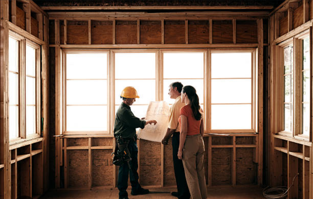 Construction worker in yellow hard hat showing blueprint to couple inside a house under construction.
