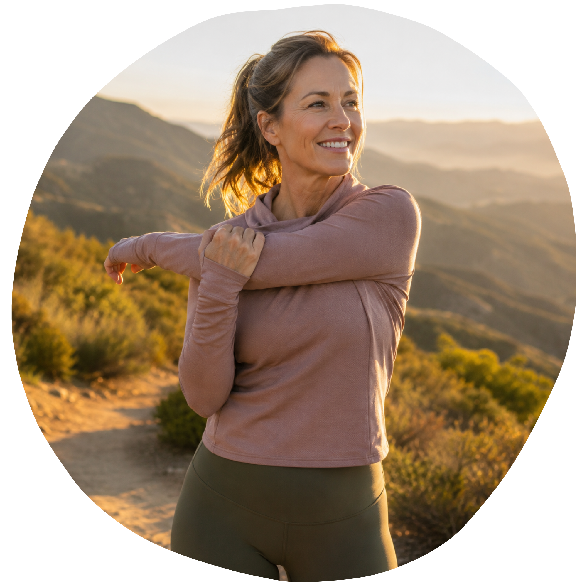 A woman stretching outdoors at sunset, wearing a mauve long-sleeve top and dark green leggings, standing on a trail with hills and trees in the background.