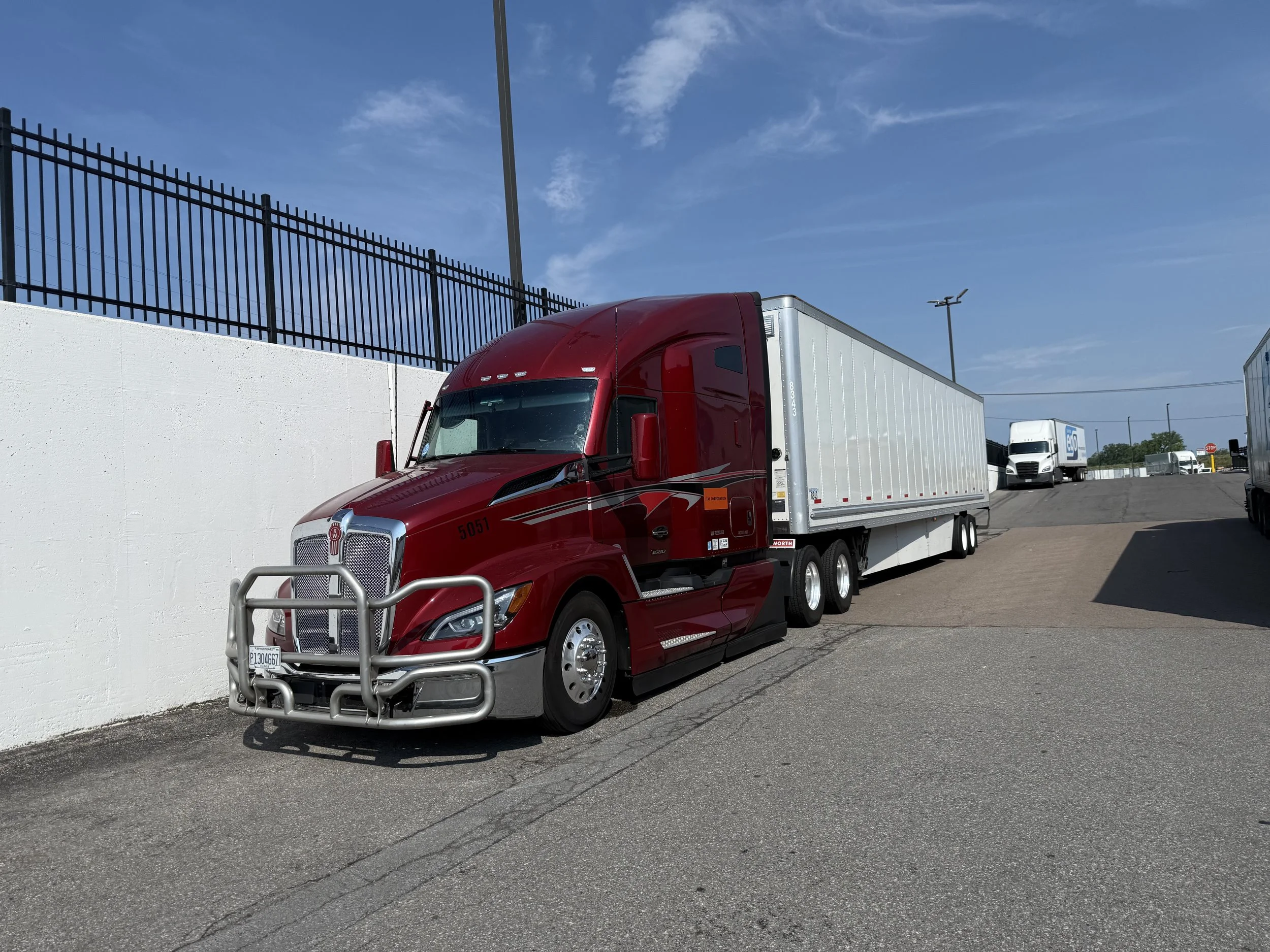 A red semi-truck with a white trailer parked near a white wall and a black fence. There are other trucks visible in the background under a blue sky with a few clouds.