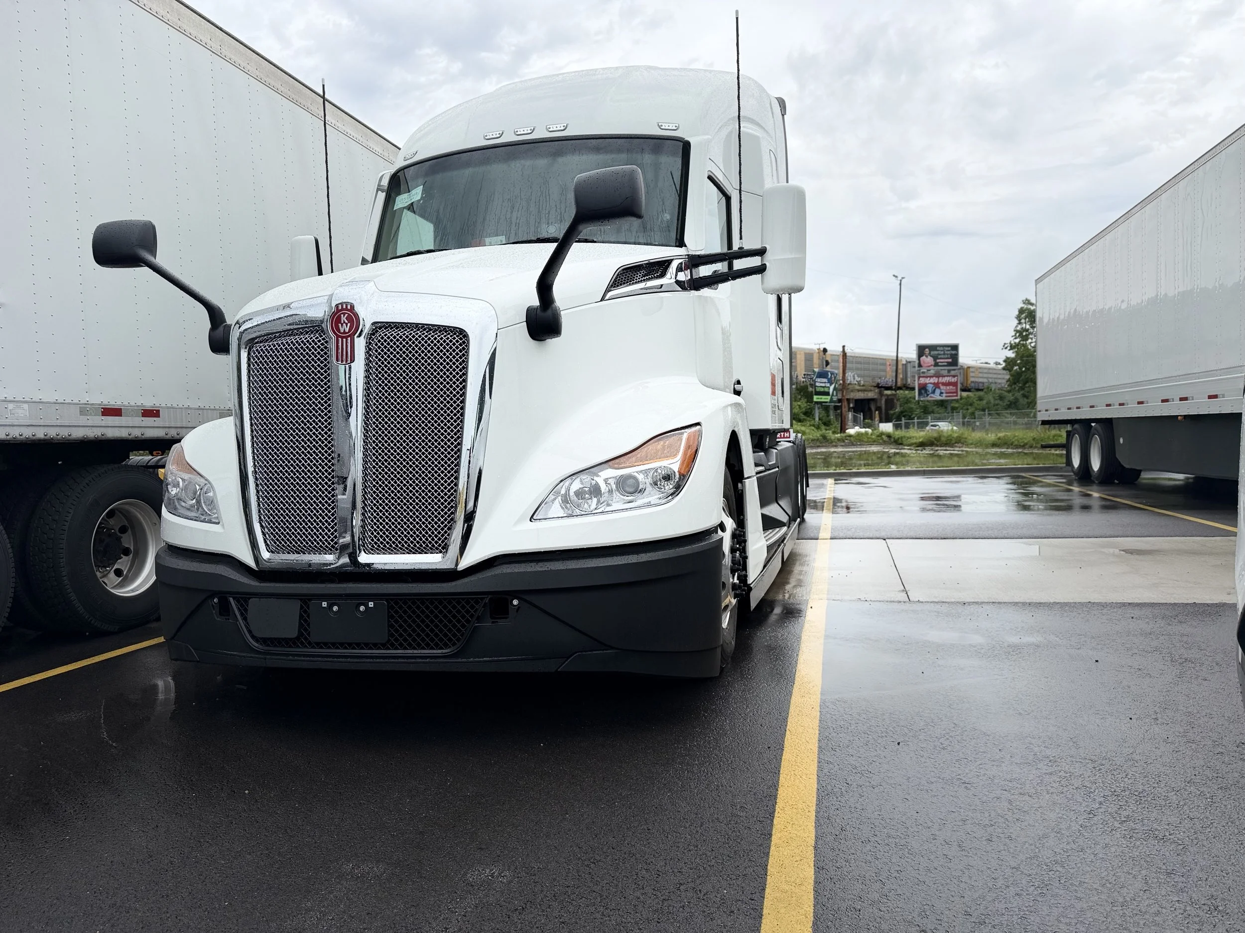 A white semi-truck parked in a lot on a rainy day, with trucks on either side and a billboard in the background.