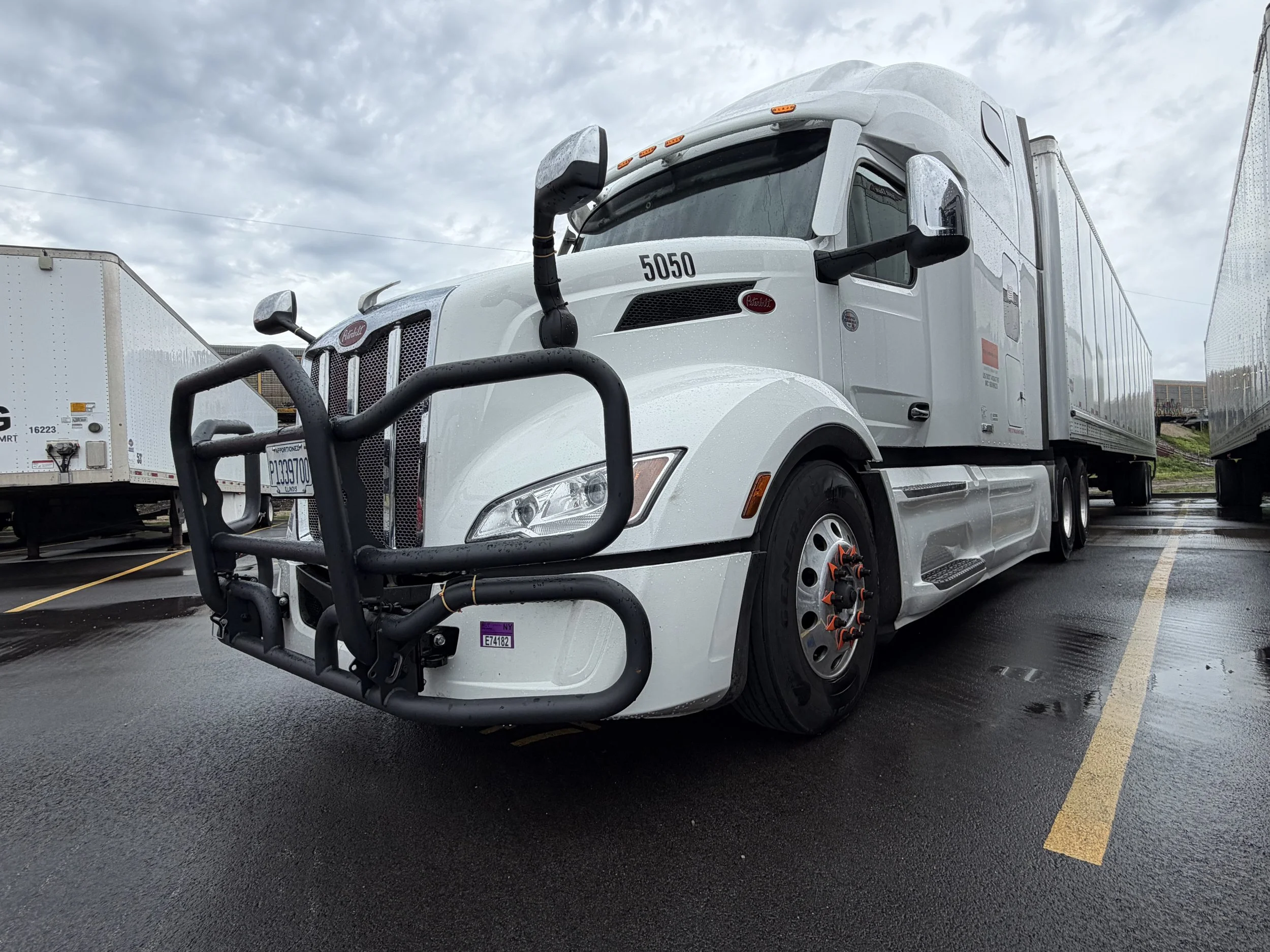 A white semi-truck with a black front bumper on wet asphalt with a cloudy sky above.