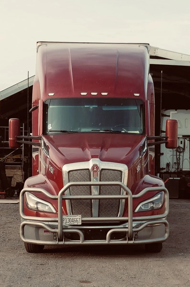 Front view of a red semi-truck with a large metal grille guard, parked at a truck stop.