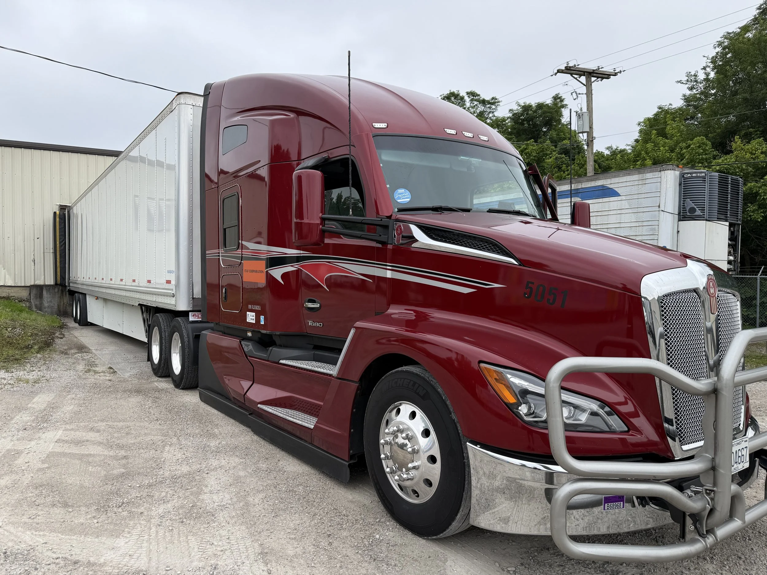A shiny red semi-truck with a white trailer parked outside on a cloudy day, with trees and utility poles in the background.