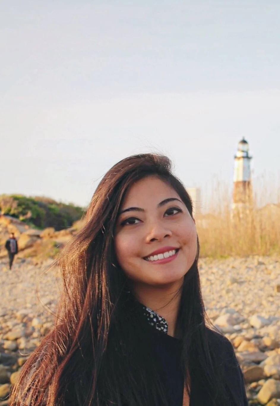 A woman smiling outdoors near a lighthouse in the distance, with rocks and dry grass around her during sunset.