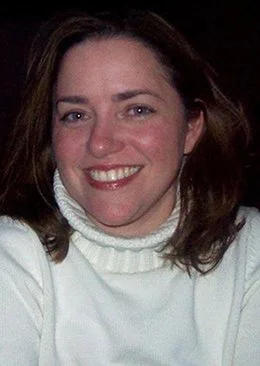 A woman smiling at the camera, wearing a white turtleneck sweater, with shoulder-length brown hair.