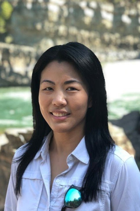 A woman with long black hair wearing a white shirt, with sunglasses tucked into her shirt, standing outdoors near rocks and water.