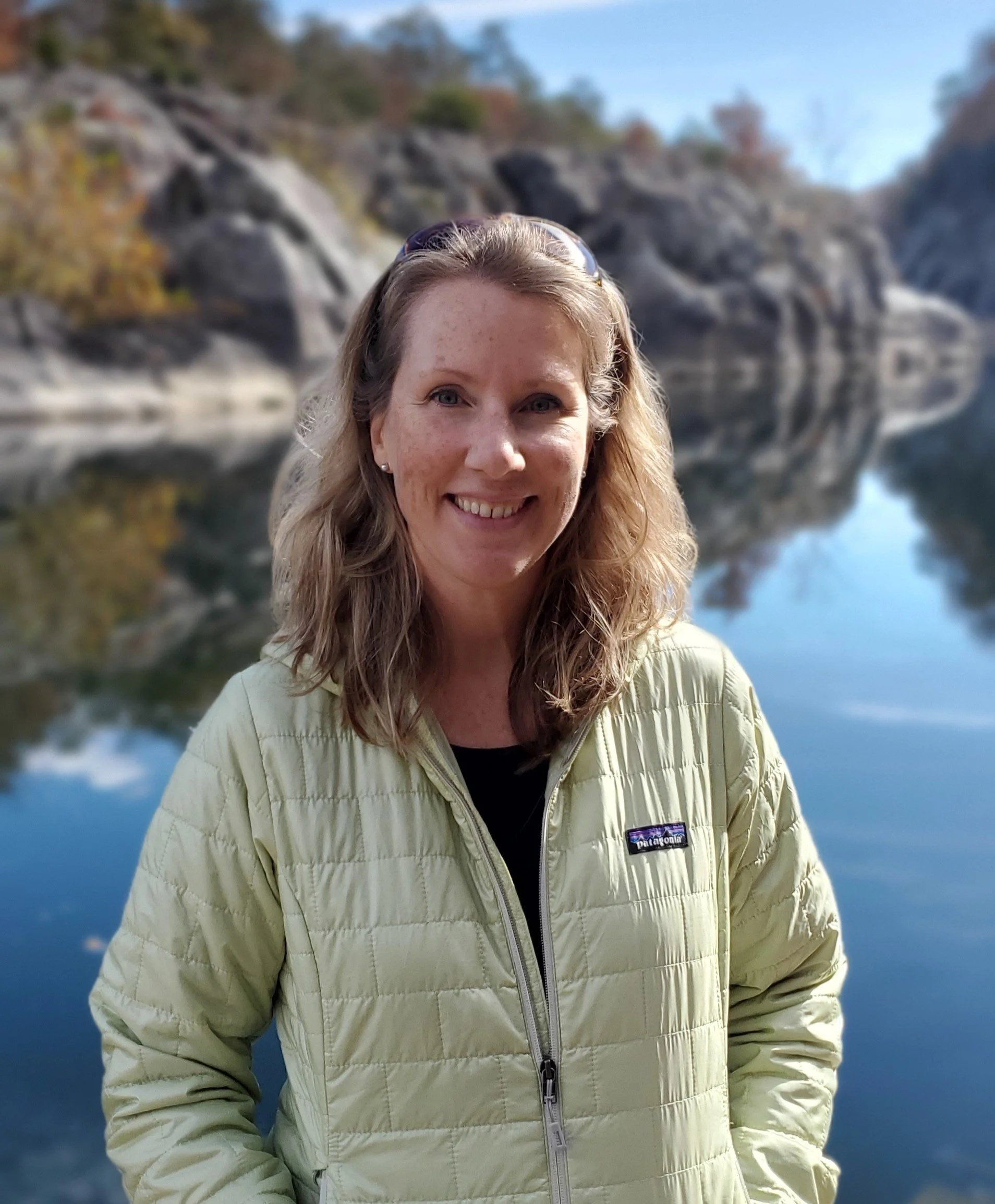 A smiling woman with blonde hair wearing a beige jacket standing outdoors near a lake or river with rocky cliffs in the background.