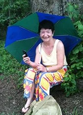 Woman sitting outdoors on a bench under a colorful umbrella, smiling and holding a smartphone.