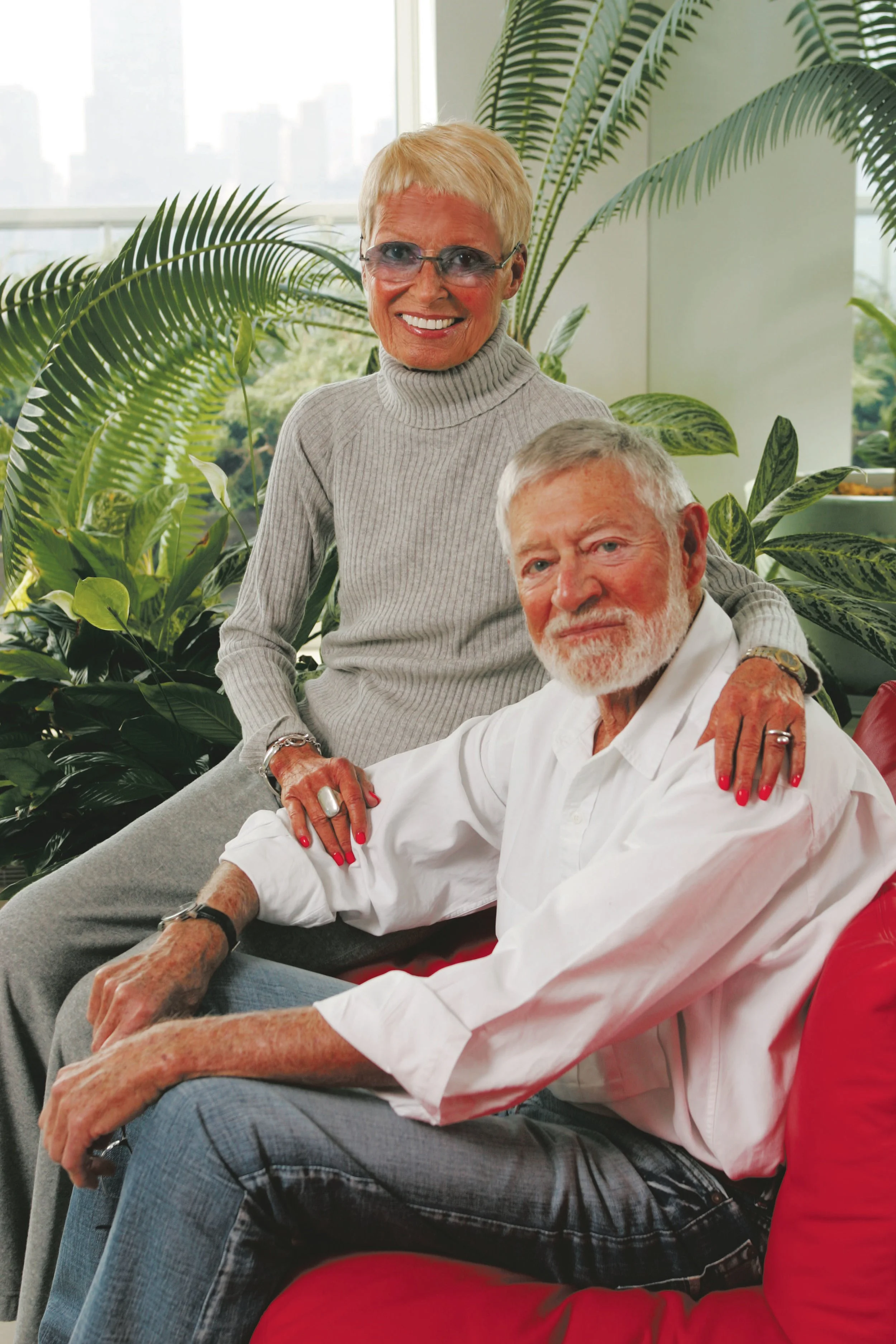 Two elderly people, a woman and a man, sitting in an indoor space surrounded by large green plants, with a cityscape visible through the windows behind them. The woman is standing and smiling at the camera; the man is seated, also smiling, with his arm resting on the woman's shoulder.