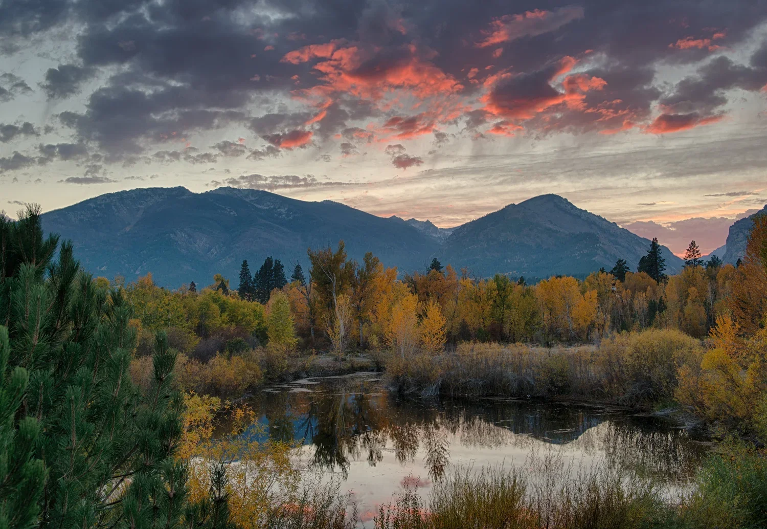 A scenic landscape featuring mountains in the background, a cloudy sky with pinkish hues, a river in the foreground, and trees with fall foliage.