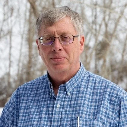 A middle-aged man with gray hair and glasses, wearing a blue checkered shirt, standing outdoors with leafless trees in the background.