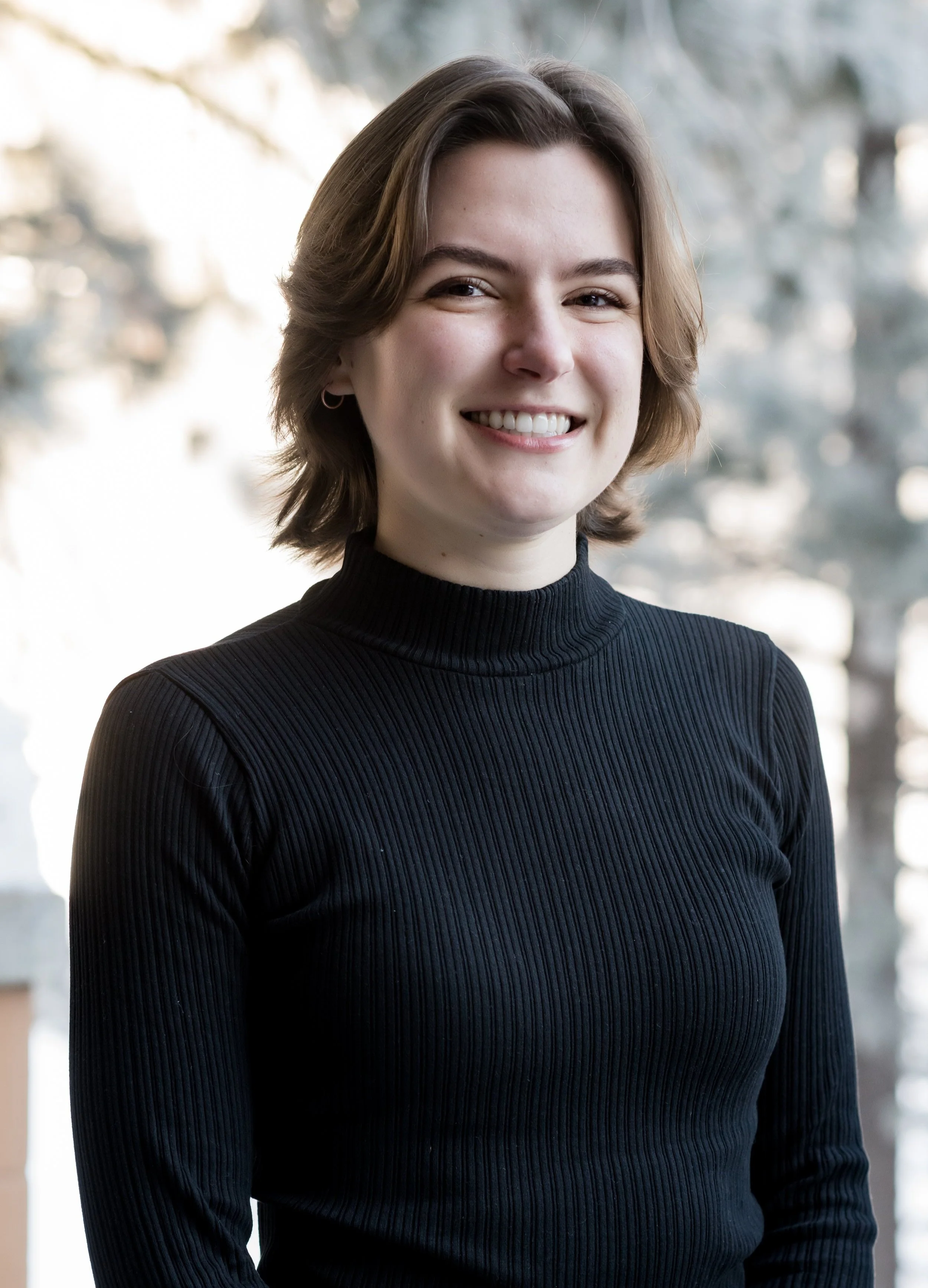 Close-up of a young woman with short brown hair, wearing a black ribbed turtleneck sweater, smiling outdoors with blurred trees in the background.