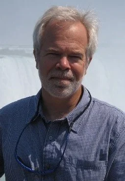A man with gray hair and a beard standing outdoors, wearing a blue checkered shirt.