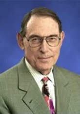 A middle-aged man wearing glasses, a light green blazer, a white shirt, and a colorful tie, standing against a blue background.