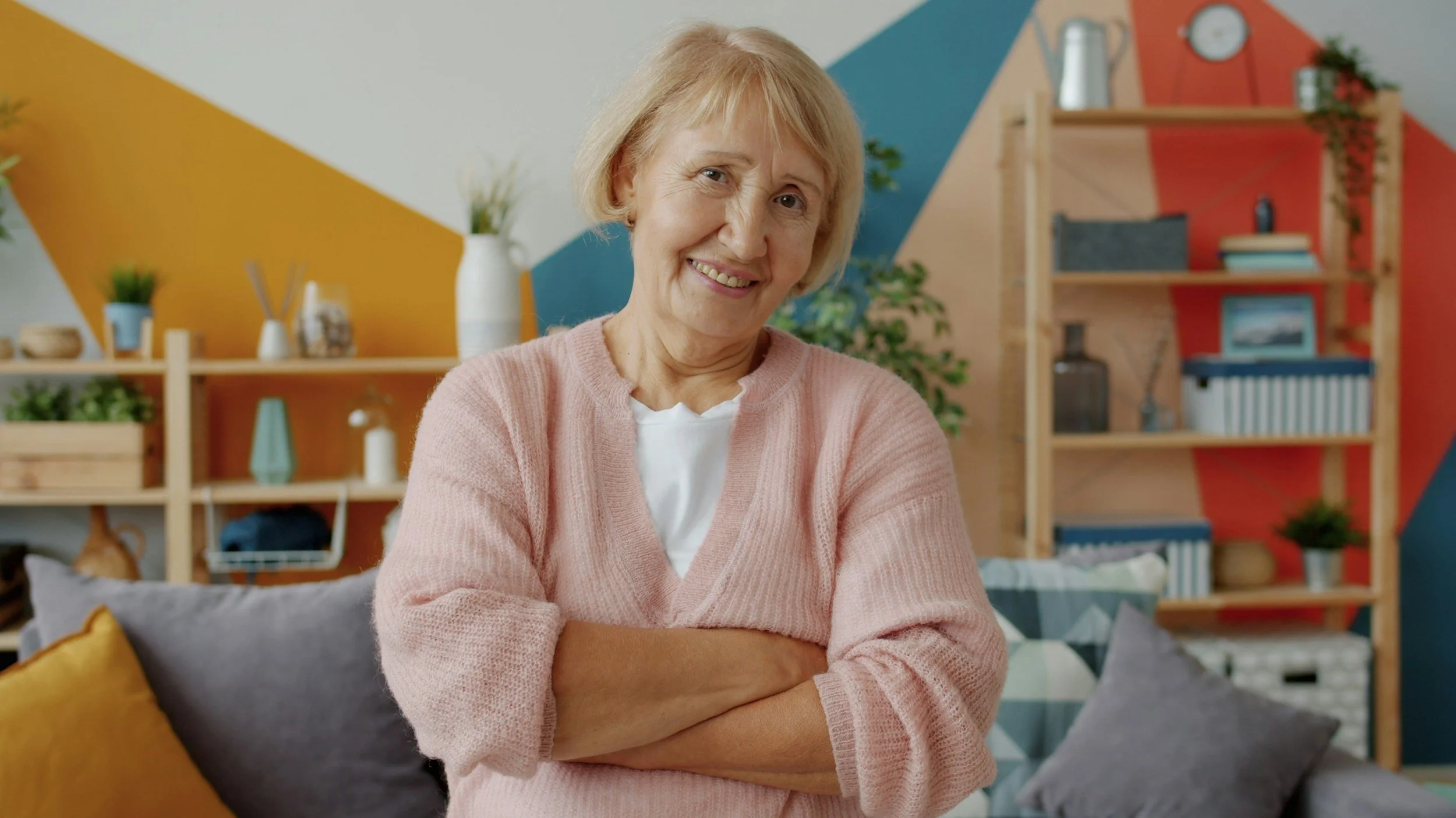 Smiling elderly woman with light blonde hair wearing a pink sweater, sitting with arms crossed in a colorful, modern living room with shelves, plants, and decorative objects.
