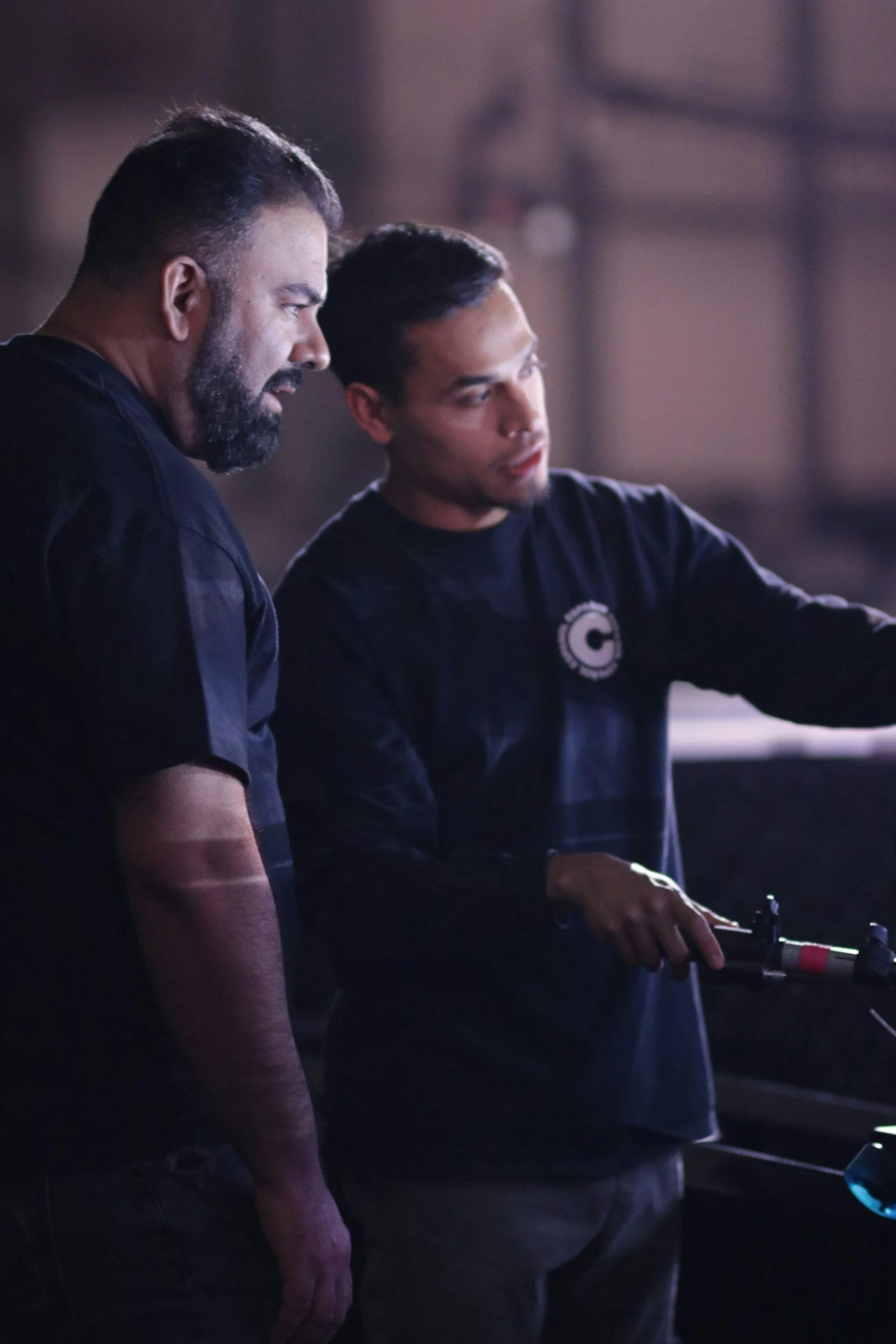 Two men are standing close together, looking at something to their right. One has a beard and short hair, the other has short dark hair. They are in a dimly lit environment, possibly a workshop or studio. The man with short dark hair is wearing a black shirt with a logo on it and appears to be manipulating some equipment.